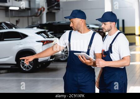 beau mécanicien en uniforme tenant une tablette numérique et pointant du doigt vers la voiture près d'un collègue dans l'atelier Banque D'Images