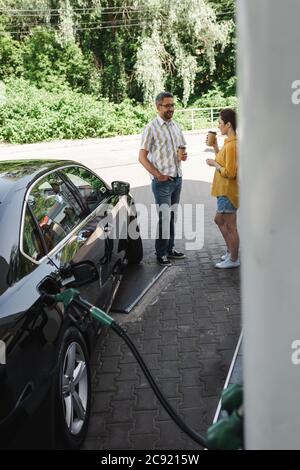 Attention sélective à l'homme souriant tenant le café pour aller près de la femme tout en faisant le plein de voiture sur la station-service Banque D'Images