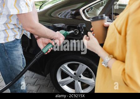 Vue courte d'un homme tenant la buse de ravitaillement près du réservoir de carburant de la voiture près d'une femme avec une tasse en papier sur la station-service Banque D'Images