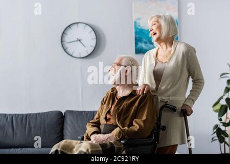 femme âgée et mari handicapé en fauteuil roulant riant tout en regardant loin Banque D'Images