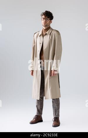 vue en longueur de l'homme branché dans un trench coat regardant la caméra tout en posant sur le gris Banque D'Images
