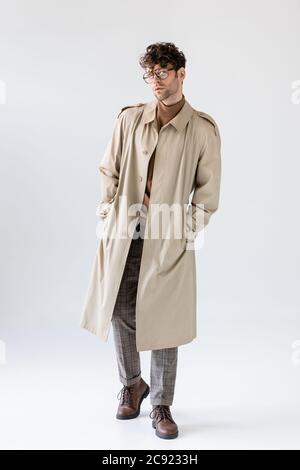 vue sur toute la longueur de l'homme tendance dans un trench-coat tenant les mains dans les poches tout en posant sur le gris Banque D'Images