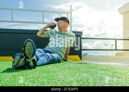 L'homme est assis sur le toit du bâtiment avec une tasse de papier de café et de téléphone cellulaire dans un T-shirt rayé et chapeau noir. Détendez-vous par beau temps. Banque D'Images