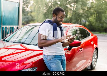 Gai décontracté hipster noir homme utilisant le smartphone et écrire des messages texte, debout près de sa voiture moderne rouge à l'extérieur Banque D'Images