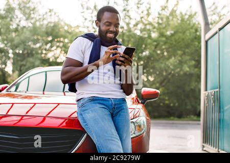 Un homme africain souriant est prêt pour un voyage en voiture. Beau jeune dar homme a la peau à l'aide de l'application de téléphone mobile ou de SMS et sourire pendant Banque D'Images