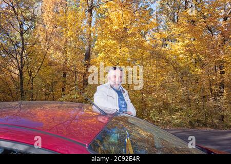 Un homme d'âge mûr repose sur le toit d'une voiture garée sur le côté d'une route forestière. Arrière-plan - magnifique feuillage jaune d'arbres Banque D'Images