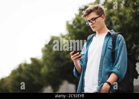 Jeune étudiant moderne de sexe masculin discutant sur le réseau en se tenant au milieu de la rue. Beau gars en lunettes avec un téléphone dans ses mains Banque D'Images