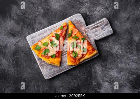 Tranches de pizza chaudes avec fromage mozzarella, jambon, tomate et persil sur planche à découper en bois, fond en béton de pierre, vue du dessus Banque D'Images