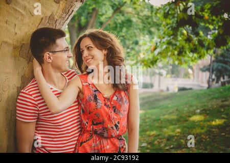 Beau couple amoureux heureux en vêtements rouges sur la nature sous un grand arbre embrassant et regardant l'un l'autre Banque D'Images