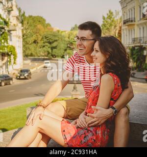 Mignon jeune couple souriant dans l'amour riant embrassant, assis à l'extérieur dans la rue verte de la ville, l'été Banque D'Images