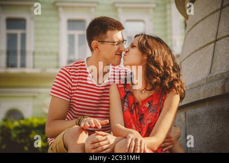 Mignon jeune couple souriant dans l'amour touchant nez, embrassant, assis à l'extérieur dans la rue verte de la ville, l'été Banque D'Images