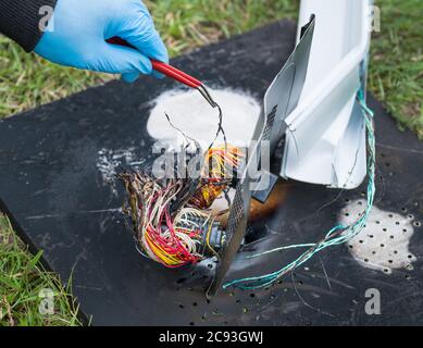 Main dans un gant de protection tenant les fils électriques endommagés avec une pince à épiler. Travail de détective sur l'épave charbrée dans l'herbe. Enquête sur une collision routière, ferroviaire ou aérienne. Banque D'Images