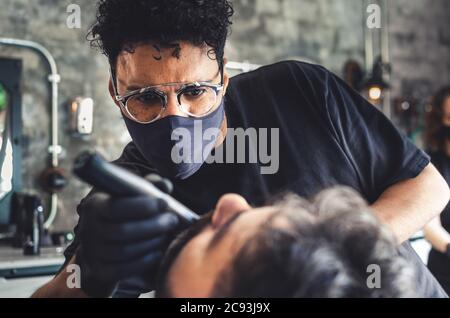 Coiffeur avec masque de protection et gants noirs pour tondre une barbe avec une tondeuse électrique Banque D'Images