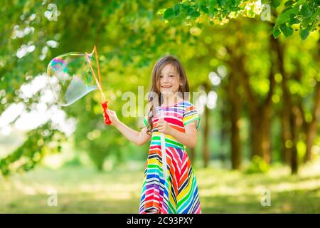 Bonne petite fille brune joue avec des bulles de savon d'été dans le parc. Mode de vie des enfants Banque D'Images