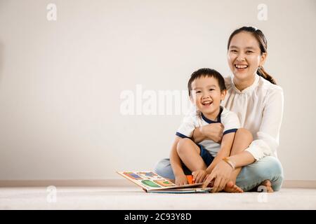 Concept de livre de lecture asiatique mère et enfant ensemble. Mère et fils vietnamiens assis sur le sol, en riant tout en retournant les pages d'un livre d'histoire, bg blanc Banque D'Images