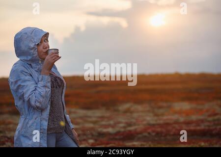 Une fille voyageur boit du thé de thermos Cup, à l'extérieur. Jeune femme buvant à la tasse. Voyage à thème. Coucher de soleil dans la toundra d'automne. Banque D'Images