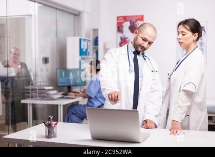 Jeunes médecins montrant l'écran d'un ordinateur portable dans le bureau de l'hôpital habillé d'un manteau blanc. Une femme âgée et un membre du couloir de la clinique parlant de prescription de la maladie. Infirmière en uniforme bleu travaillant sur l'ordinateur. Banque D'Images