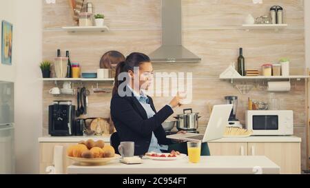 Femme d'affaires agitant sur appel vidéo pendant le petit déjeuner. Jeune indépendant dans la cuisine ayant un repas sain tout en parlant sur un appel vidéo avec ses collègues du bureau, en utilisant la technologie moderne et de travail 24 heures sur 24 Banque D'Images