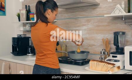 Femme craquant les œufs dans la poêle pour le petit déjeuner. Une matinée saine avec des produits frais, un mode de vie heureux pour la femme au foyer qui cuisine dans une cuisine moderne et confortable sous la lumière chaude et ensoleillée du matin d'été Banque D'Images