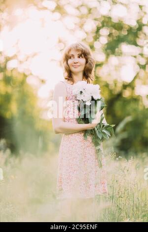 Belle jeune femme blonde caucasienne en robe légère tenant un bouquet de pivoines blanches, marchant dans le champ d'été ou jardin au coucher du soleil. Femme avec fleurs Banque D'Images