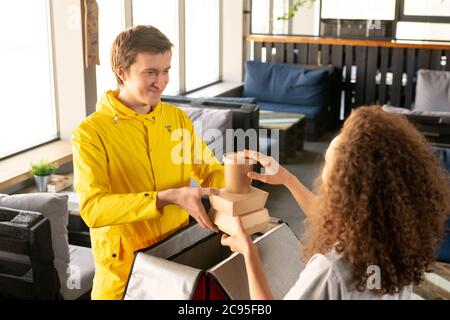 Jeune coursier souriant en blouson jaune emballant les boîtes alimentaires dans un sac thermique avec serveuse dans un café vide pendant l'épidémie de coronavirus Banque D'Images