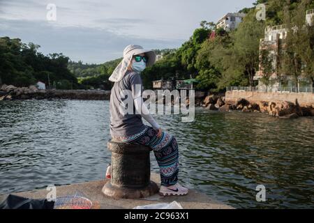 Une femme portant un masque facial a vu la pêche à la jetée du ferry à Yung Shue WAN, sur l'île de Lamma. L'été 2020 est marqué par les scènes liées à la pandémie du coronavirus. Masques chirurgicaux et avertissements interdisant aux personnes d'accéder au scénario. Banque D'Images