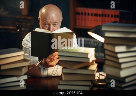 Un écrivain âgé lit à table avec une pile de livres Banque D'Images