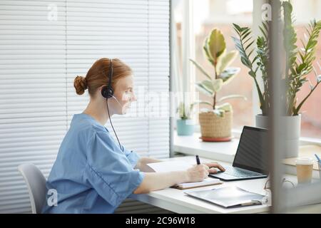 Femme médecin dans un casque assis à la table devant l'ordinateur portable et prenant des notes dans le bloc-notes pendant son travail Banque D'Images