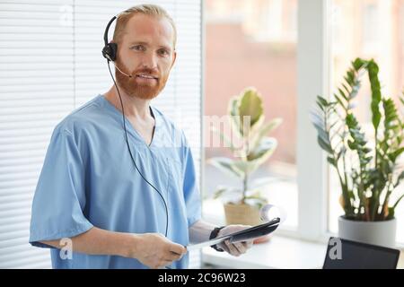 Portrait d'un opérateur médical masculin en uniforme et casque souriant à la caméra tout en se tenant au bureau avec des documents Banque D'Images