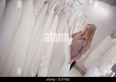 Une jeune femme blonde choisit une robe de mariage dans un salon de mariage. La mariée-à-être est à la recherche de la robe parfaite pour elle-même. Magasin de vêtements. Banque D'Images