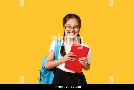 Adorable écolière chinoise avec sac à dos tenant le livre sur fond jaune Banque D'Images