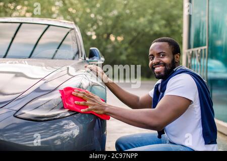 Portrait d'un jeune homme à la peau sombre souriant en tenue décontractée, nettoyant son phare de voiture électrique moderne avec un tissu en microfibre rouge à l'extérieur, à la Banque D'Images