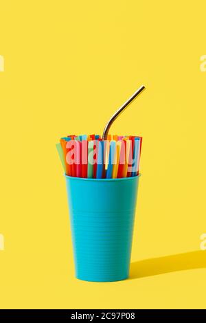 une paille réutilisable en acier inoxydable se distingue de quelques pailles jetables en plastique de différentes couleurs dans un vase bleu, sur un bac jaune Banque D'Images