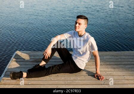 Paisible homme dans une tenue décontractée assis sur un quai en bois près du lac pendant le coucher du soleil et regardant loin Banque D'Images