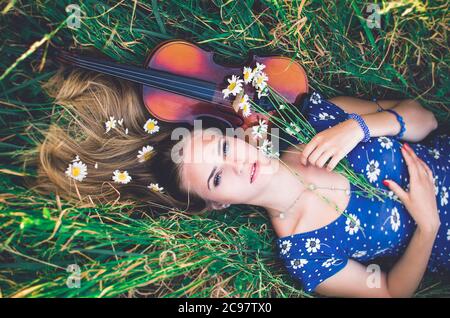 Jeune fille mince à la peau équitable blonde belle mensonges dans l'herbe haute. Vue de dessus. Fille dans une robe courte bleue avec un imprimé de marguerites. Un bouquet et lo Banque D'Images