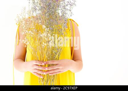 Femelle méconnaissable en robe jaune se cachant derrière un bouquet de fleurs sauvages le jour de l'été sur fond blanc Banque D'Images