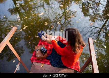 Une jeune femme est assise avec son dos sur le pont près du lac lors d'une belle journée ensoleillée, chaude et belle automne. Couverture en tissu écossais, ancien appareil photo. Banque D'Images