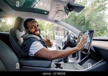 Portrait d'un jeune homme africain souriant et charmant avec une barbe assise à l'intérieur de sa voiture moderne à direction automatique et essuyant la poussière de l'écran tactile avec Banque D'Images