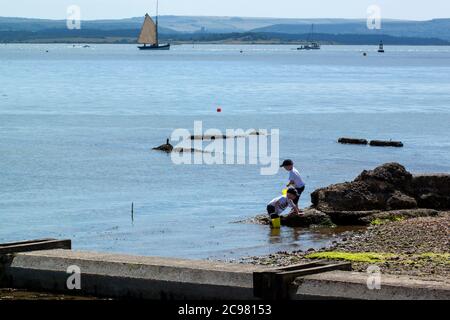 Bristol/UK 04/17/2010: Les deux garçons jouent avec des jouets de plage sur une petite plage sale et robuste recouverte de pierres de galets, de rochers et de mousse. Les bateaux sont visib Banque D'Images