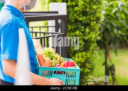 L'homme de livraison de magasin d'épicerie asiatique portant l'uniforme bleu et le masque facial protègent il livrant des légumes frais dans une boîte en plastique à la porte avant de la maison après Banque D'Images