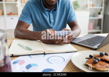 Gros plan sur un homme d'affaires africain assis à la table avec des documents et utilisant un téléphone portable dans son travail en ligne à la maison Banque D'Images