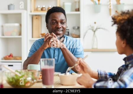 Père souriant africain parlant à son fils tout en étant assis à la table pendant le petit déjeuner à la maison Banque D'Images