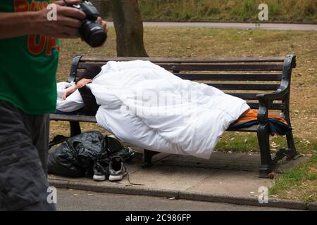 Un homme anonyme et un homme sans abri non identifiable dormait sur un banc de parc dans un espace public d'une ville côtière tandis que d'autres personnes marchent devant. Bournemouth, Angleterre, Royaume-Uni. (120) Banque D'Images