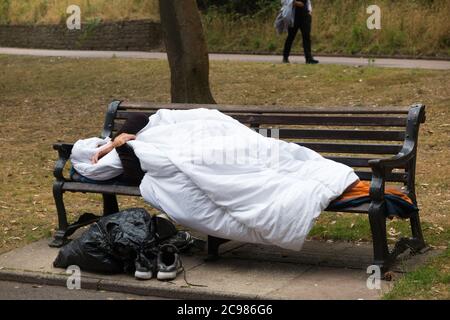 Un homme anonyme et un homme sans abri non identifiable dormait sur un banc de parc dans un espace public d'une ville côtière tandis que d'autres personnes marchent devant. Bournemouth, Angleterre, Royaume-Uni. (120) Banque D'Images