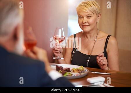 Femme de race blanche en robe noire fine nourriture de dîner avec l'homme dans un restaurant élégant. Une femme a levé un verre et va boire. Vue arrière sur mari Banque D'Images