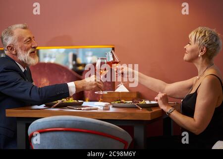 Un couple haut de gamme et élégant s'assoit au restaurant et des verres à champagne sont à l'honneur. Homme à poil gris barbu en tuxedo et femme caucasienne en DR noir Banque D'Images