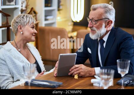 Joyeux, joyeux, heureux, couple mature amoureux de passer du temps au restaurant tout en discutant et regardant les uns les autres. Un homme âgé montre quelque chose sur une tablette Banque D'Images