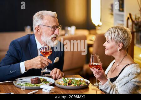 Un couple senior élégant et élégant se regarde les uns les autres et discute, entretient des relations étroites. Un homme aux cheveux gris en tuxedo lève un verre. Utilisez une cigarette électronique, un singe. Mo Banque D'Images