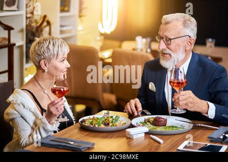 Un couple senior élégant et élégant se regarde les uns les autres et discute, entretient des relations étroites. Un homme aux cheveux gris en tuxedo lève un verre. Utilisez une cigarette électronique, un singe. Mo Banque D'Images