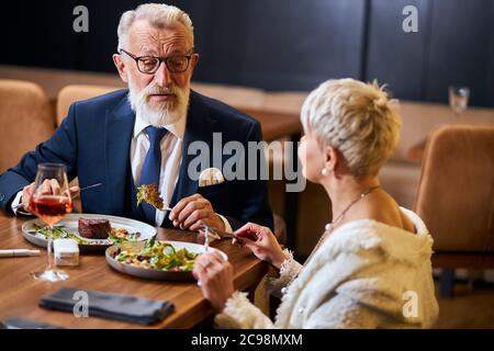 Jolie dame et homme âgé aux cheveux gris dans tuxedo ayant une conversation amicale dans le restaurant. Les collègues après le travail discutent et mangent Banque D'Images